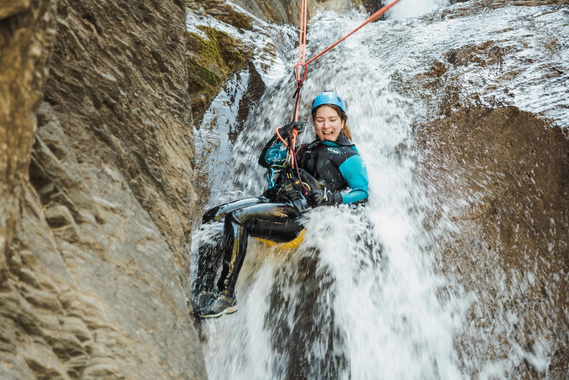 A smiling woman in a wetsuit and helmet rappels down a powerful waterfall.