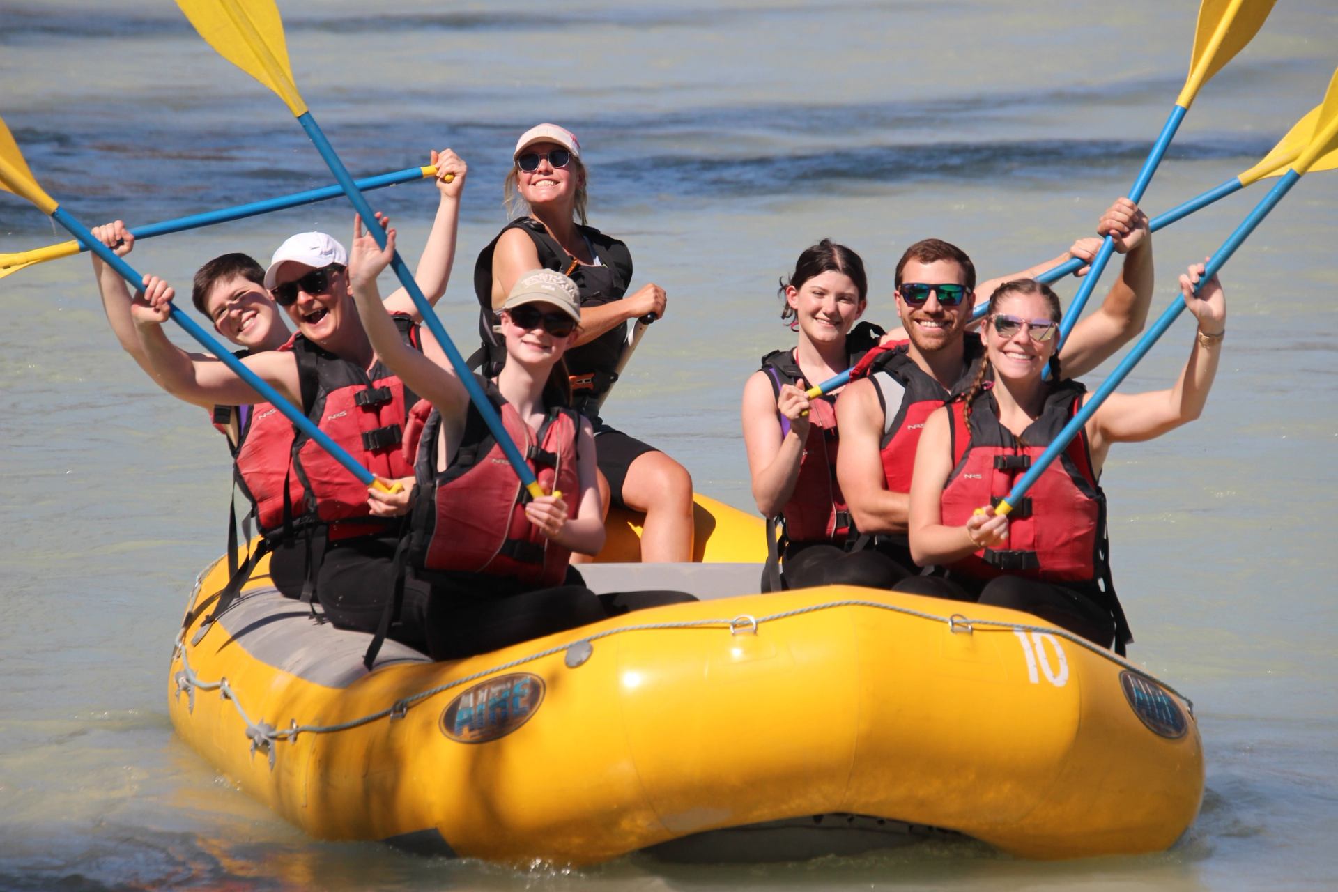 Group raising paddles while rafting on calm, light-blue water in a yellow raft.