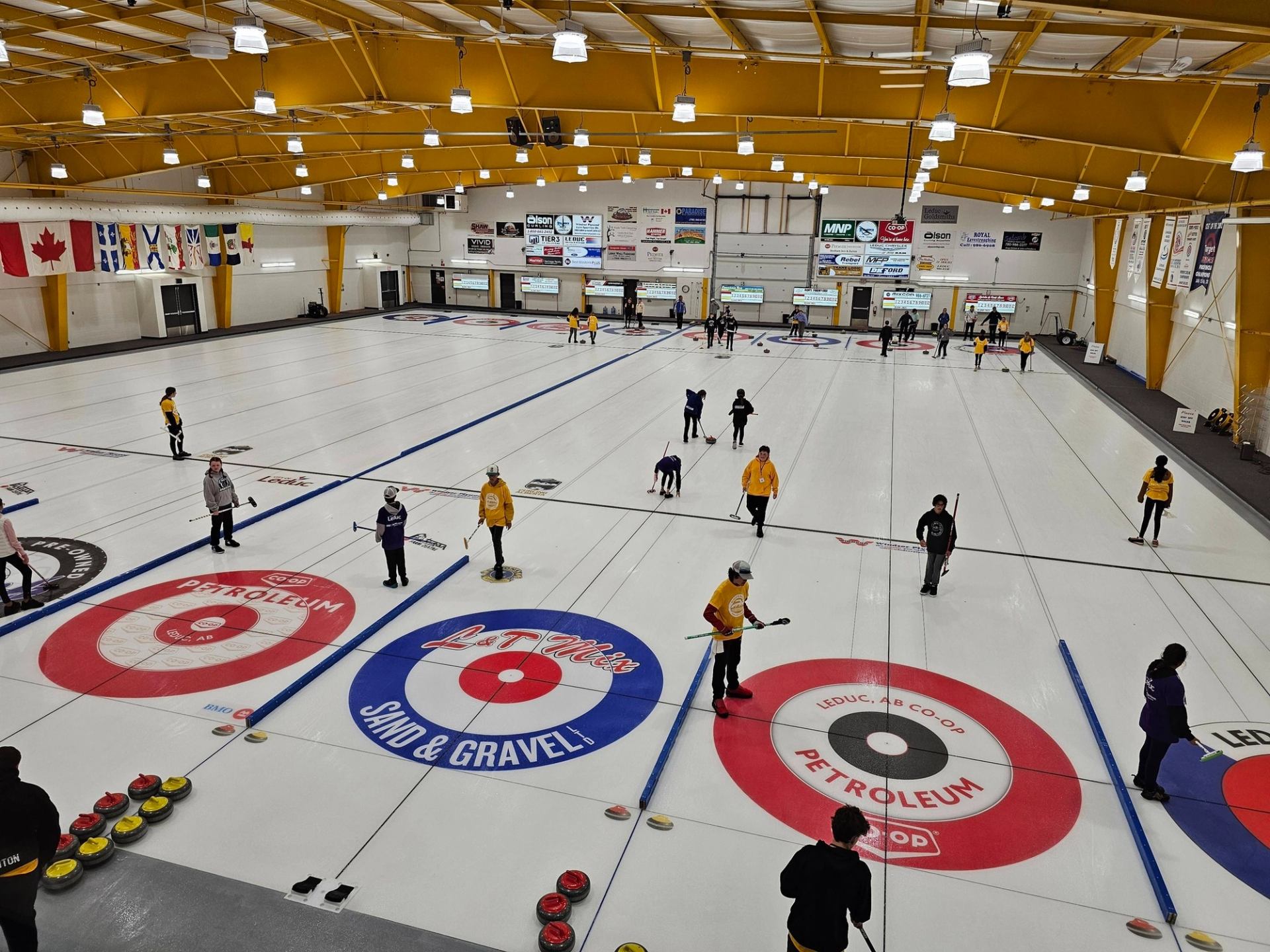 Indoor curling rink with multiple sheets, players sweeping stones, and flags hanging overhead.