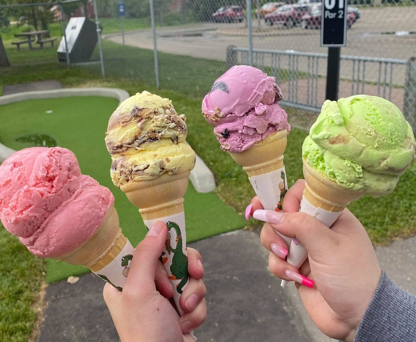 Hands holding four ice cream cones with pink, beige, and green scoops at a mini golf course.