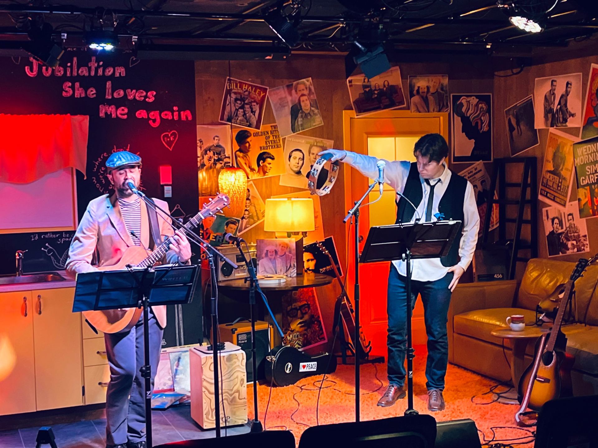 Two performers sing and play guitar on a cozy stage with posters and warm lighting.