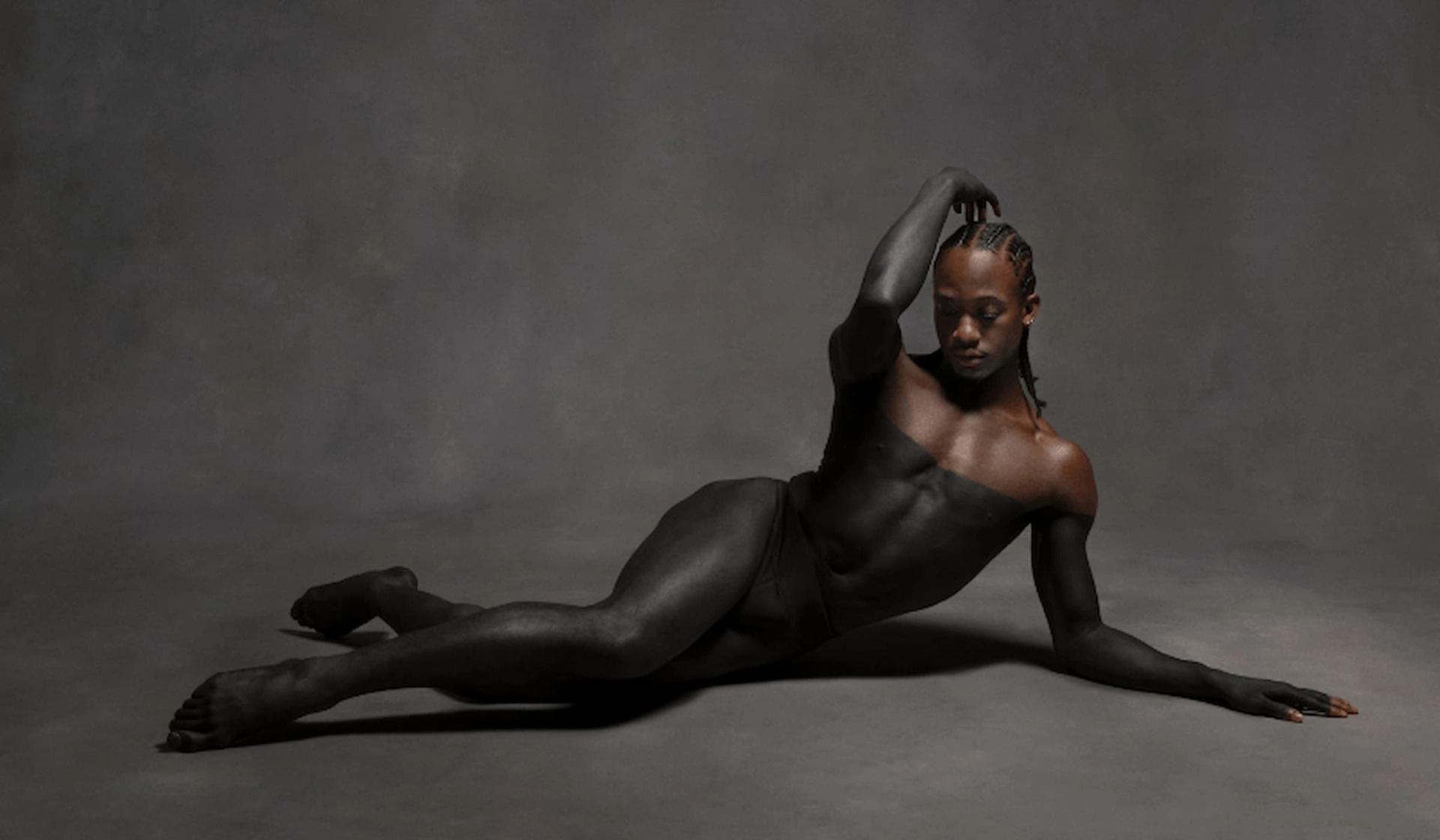 Person with braided hair lies on studio floor, body painted dark, arm raised overhead.