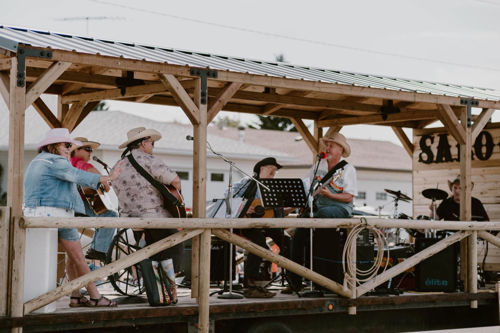 Country band performs on wooden outdoor stage with guitars, drums and microphones.