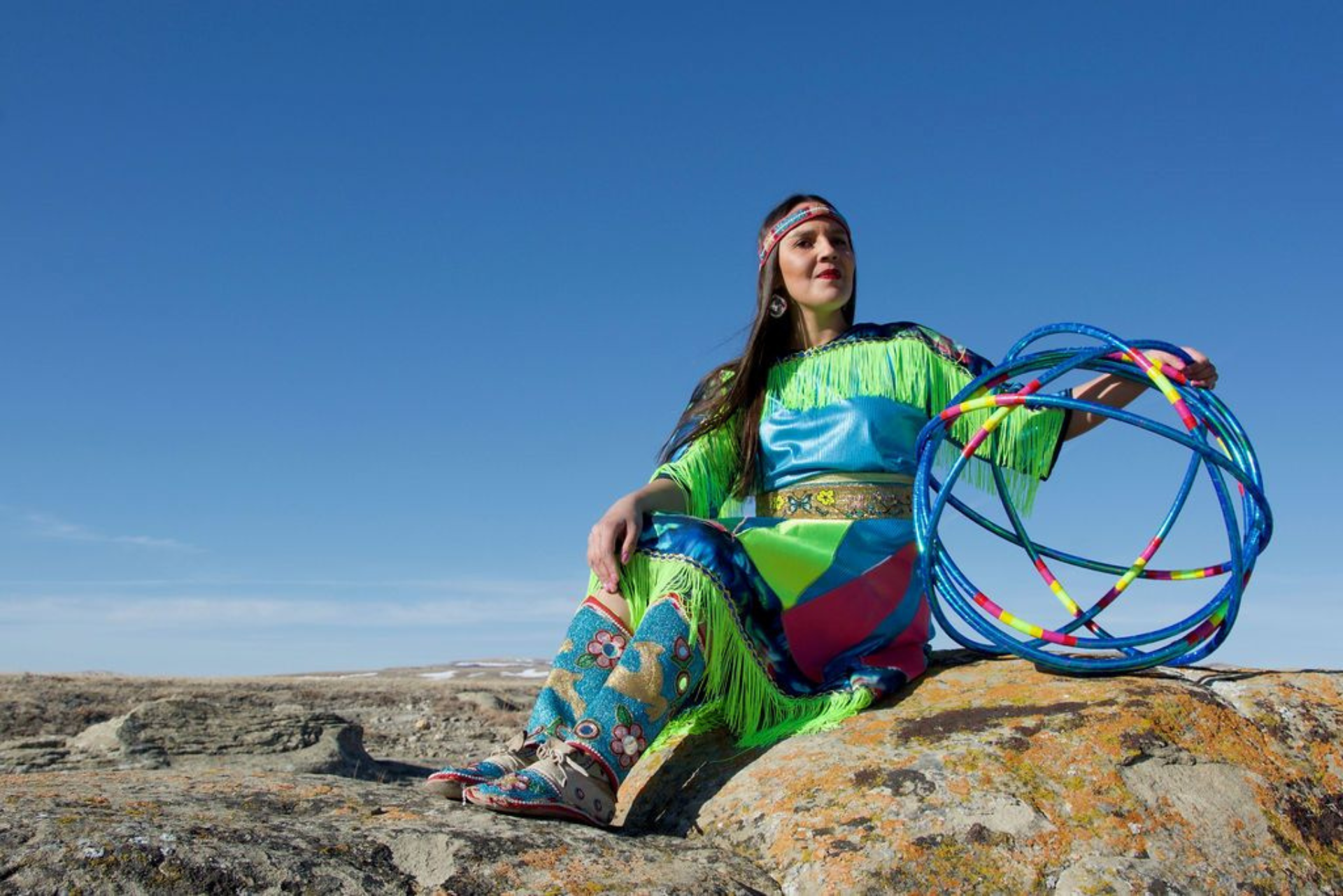 Hoop dancer in colourful regalia sits on rocky ground under clear blue sky.