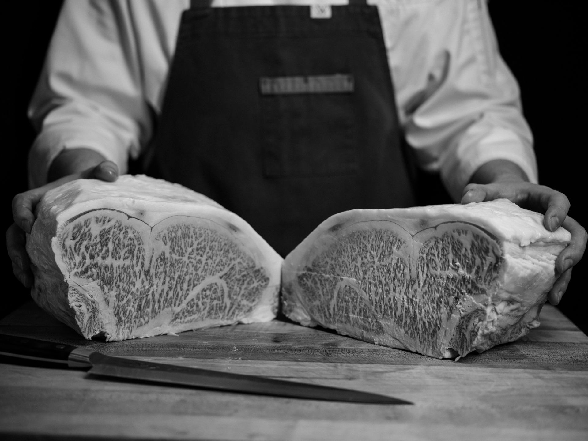 Chef holding two large marbled beef cuts on a wooden board with a knife in front.