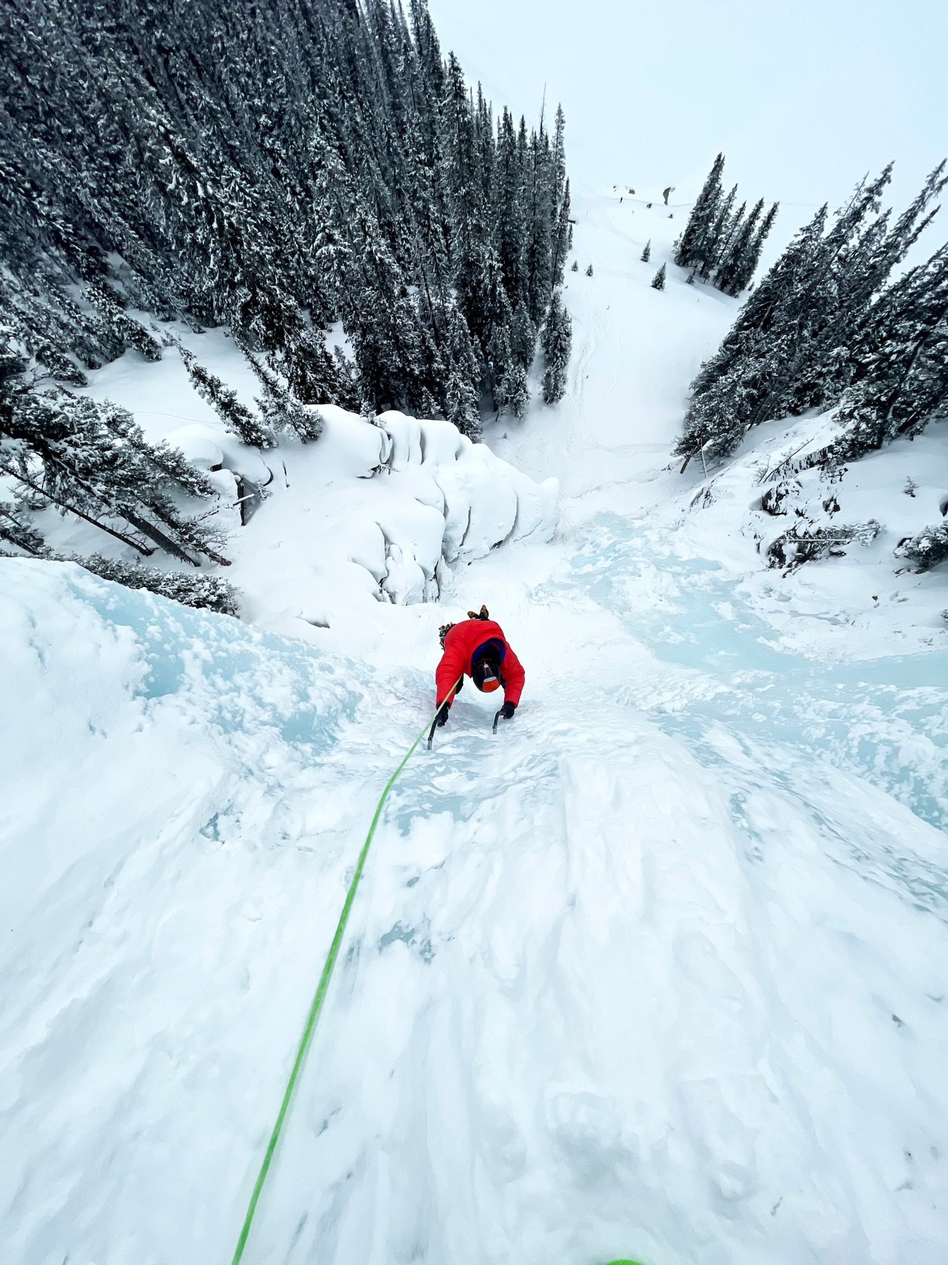 Climber ascending a steep frozen waterfall with rope, surrounded by snow-covered trees and alpine terrain.