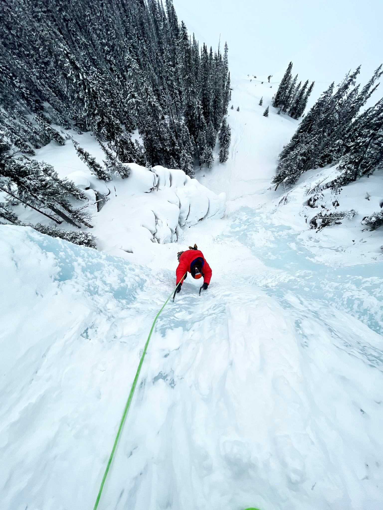 Climber ascending a steep frozen waterfall with rope, surrounded by snow-covered trees and alpine terrain.