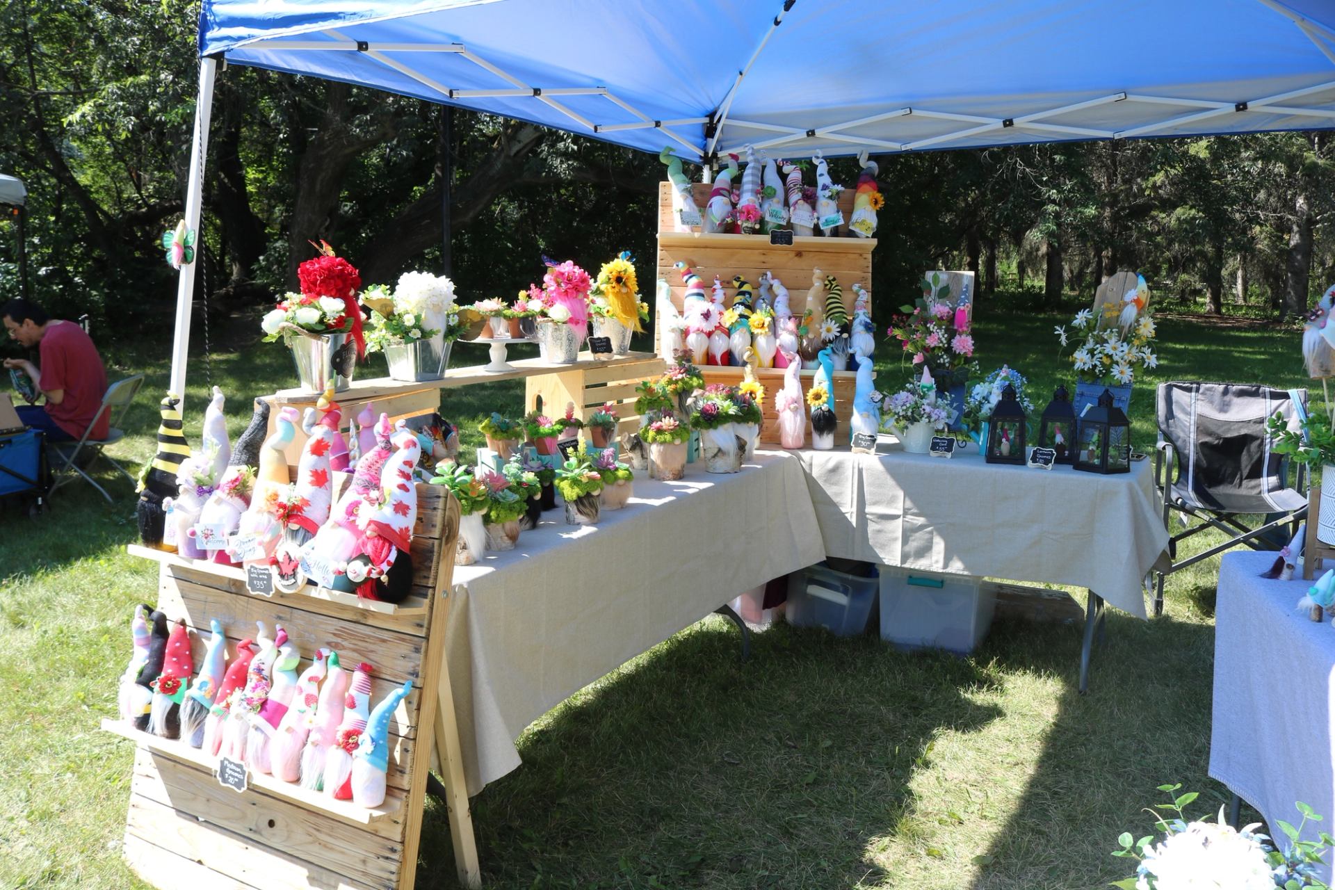 A market booth displaying handmade gnome crafts and flower arrangements outdoors.