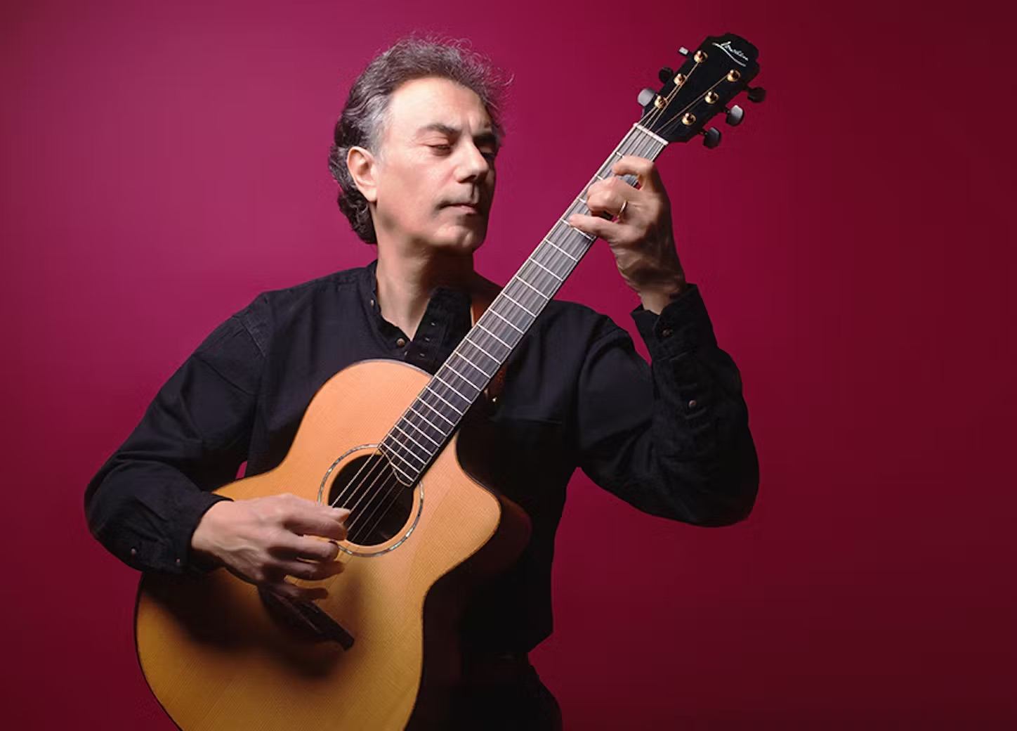 Guitar player holding acoustic guitar on maroon background, hand on fretboard and strings.