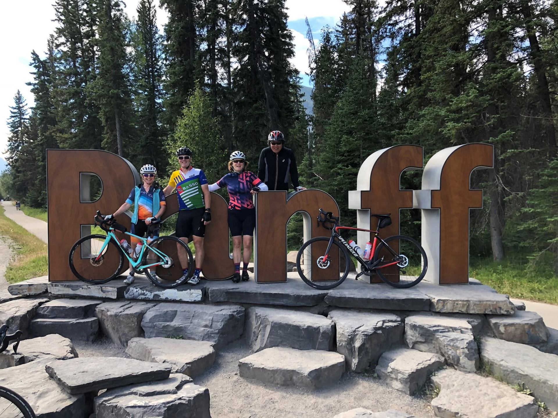 Cyclists with road bikes stand beside a Banff sign among evergreen trees.