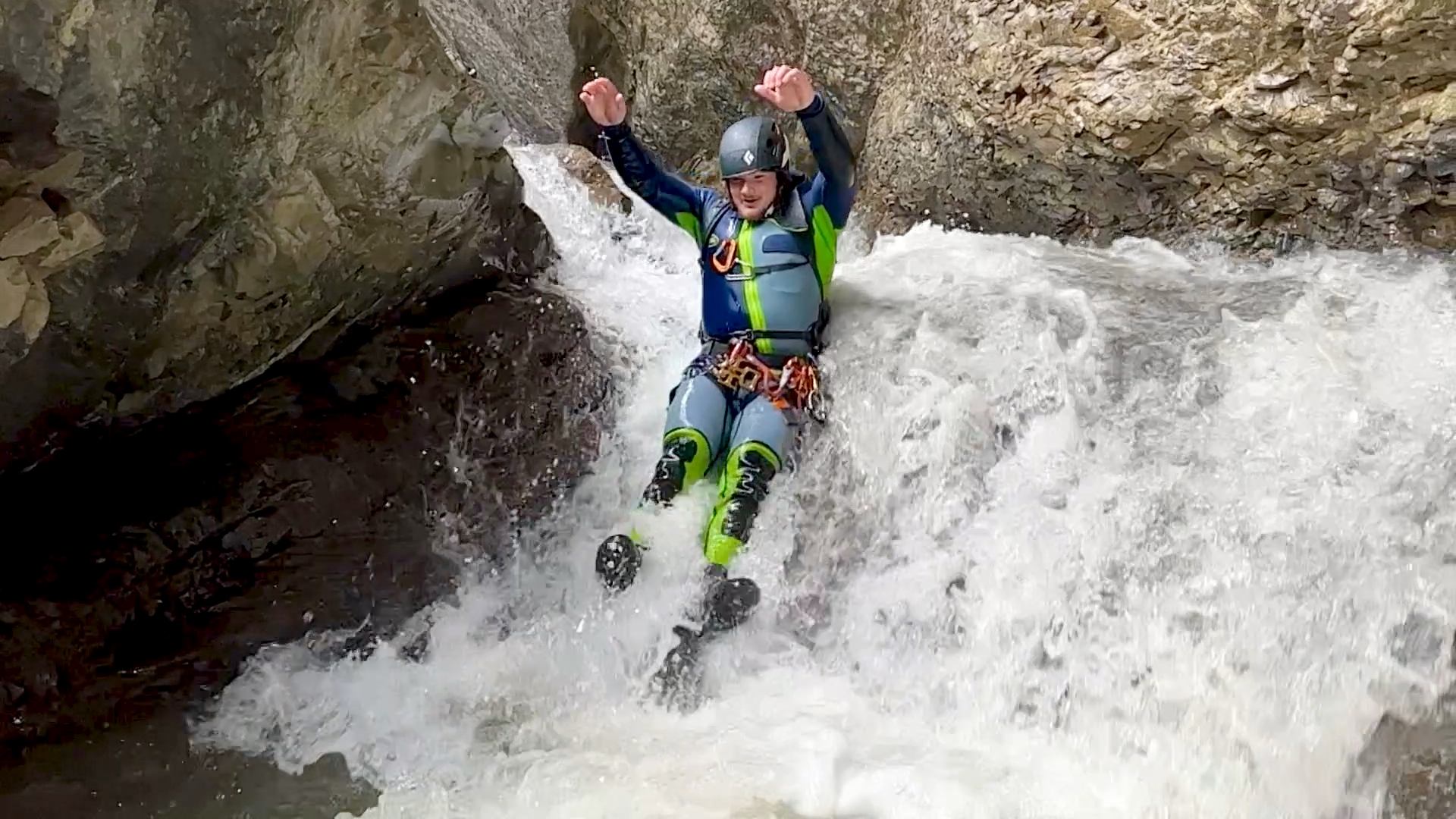 Person sliding down a rushing waterfall wearing a wetsuit and helmet.