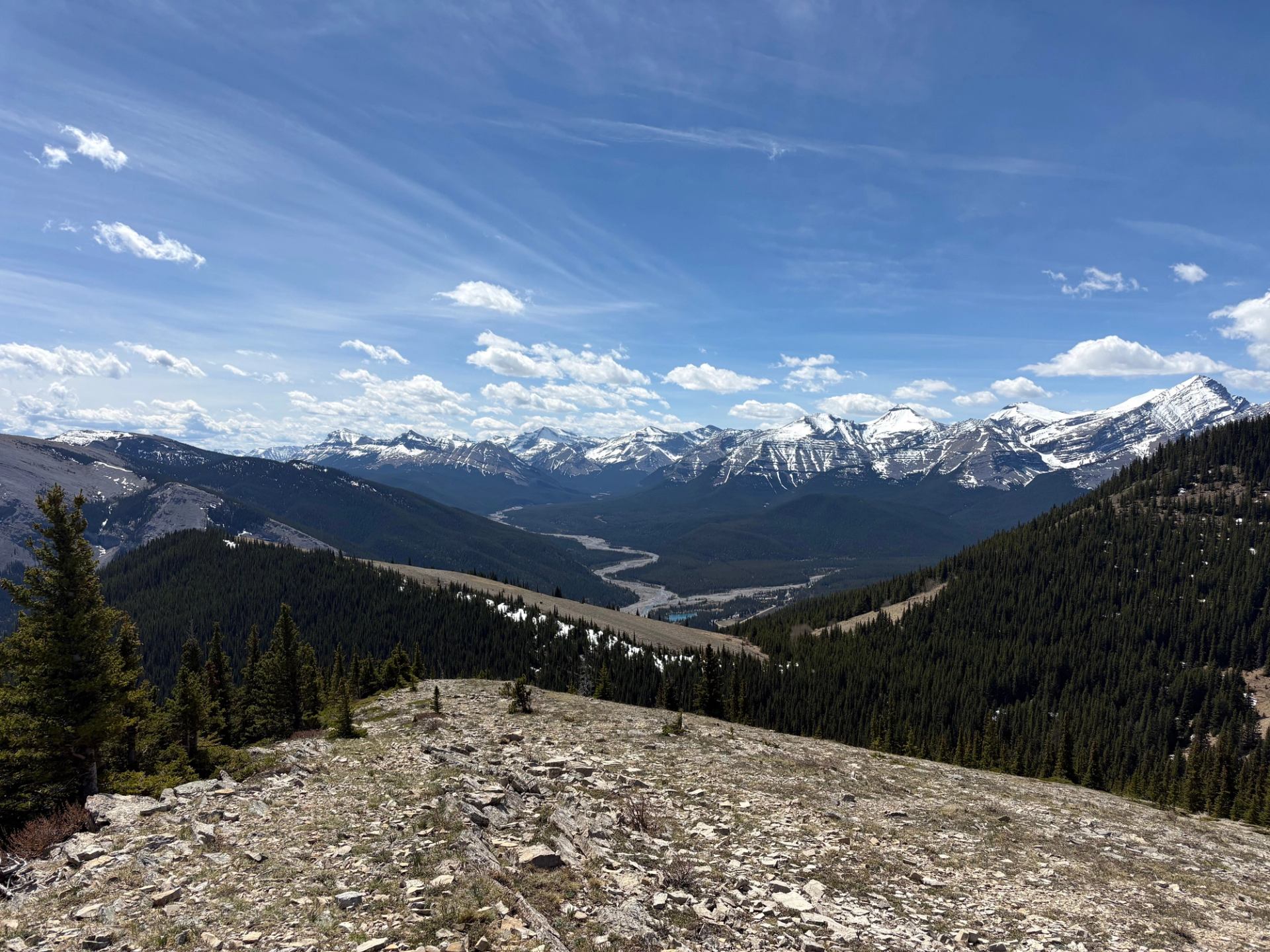 Rocky mountain ridge overlooking forested valleys, winding river and distant snow-capped peaks.