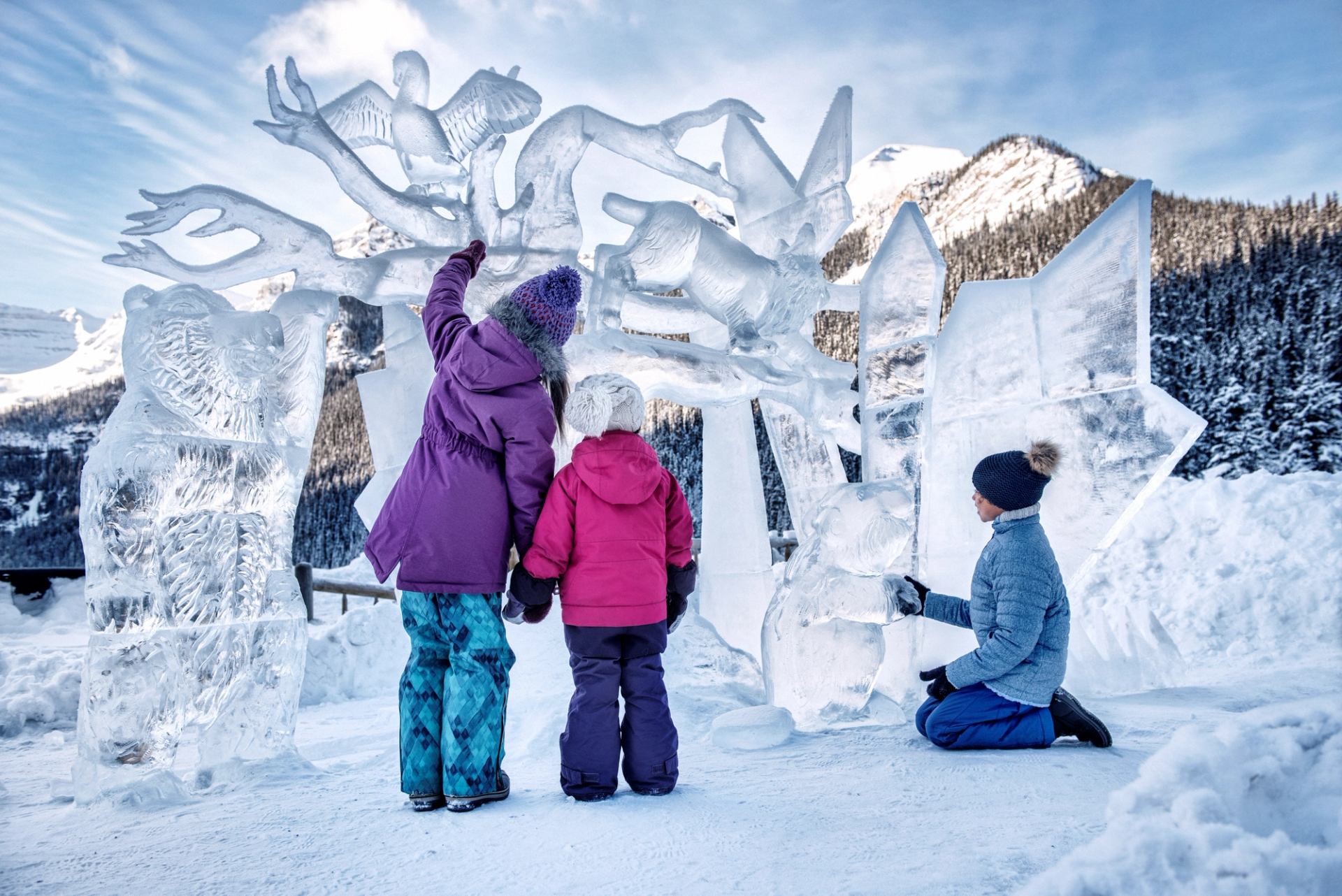 Children in winter gear pose by detailed ice sculptures with mountains behind.