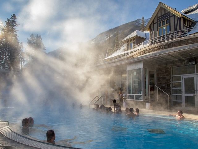 People relaxing in steaming outdoor pool with snowy mountains in the background.