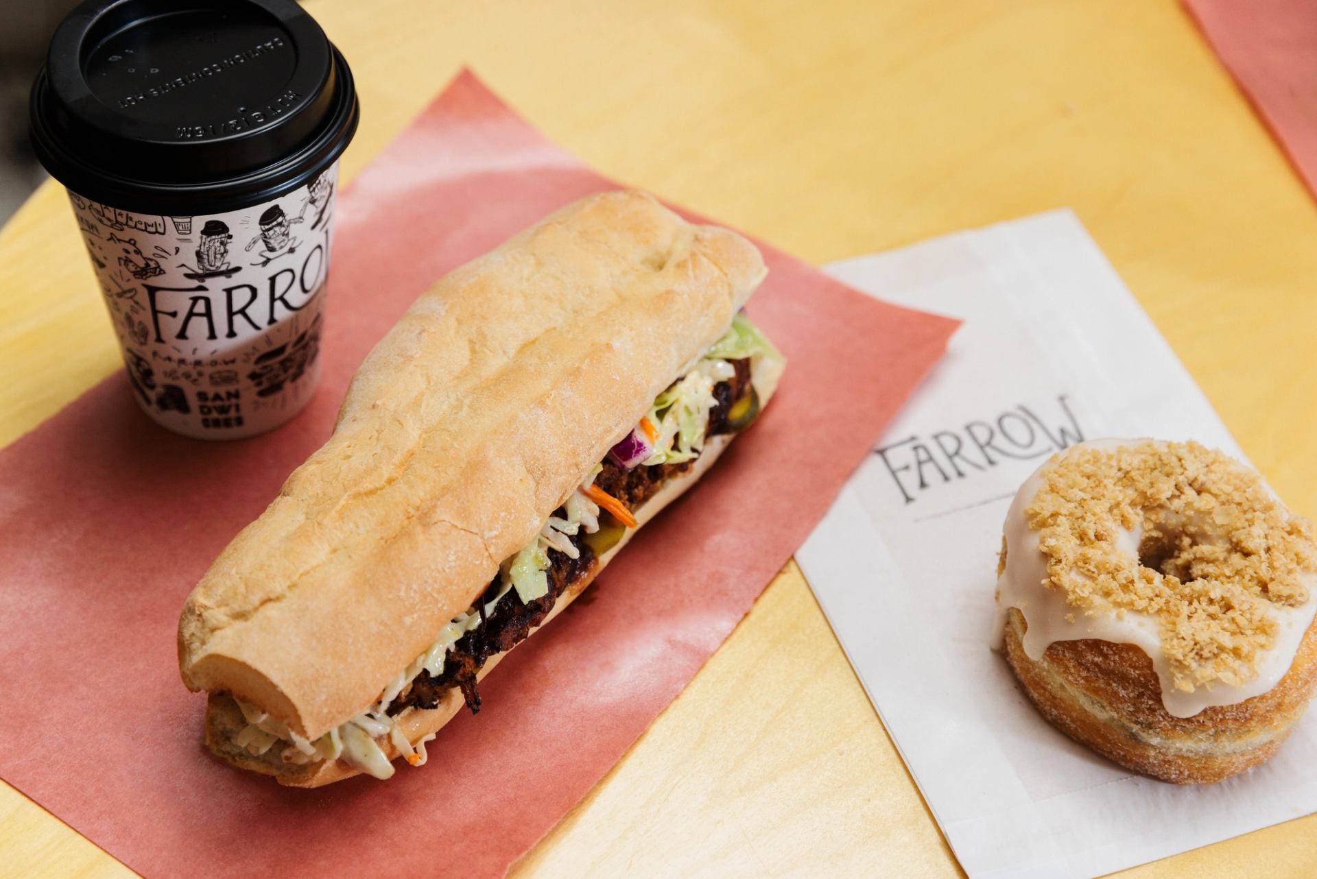 Farrow coffee cup, filled sandwich and iced doughnut sit on a yellow table.