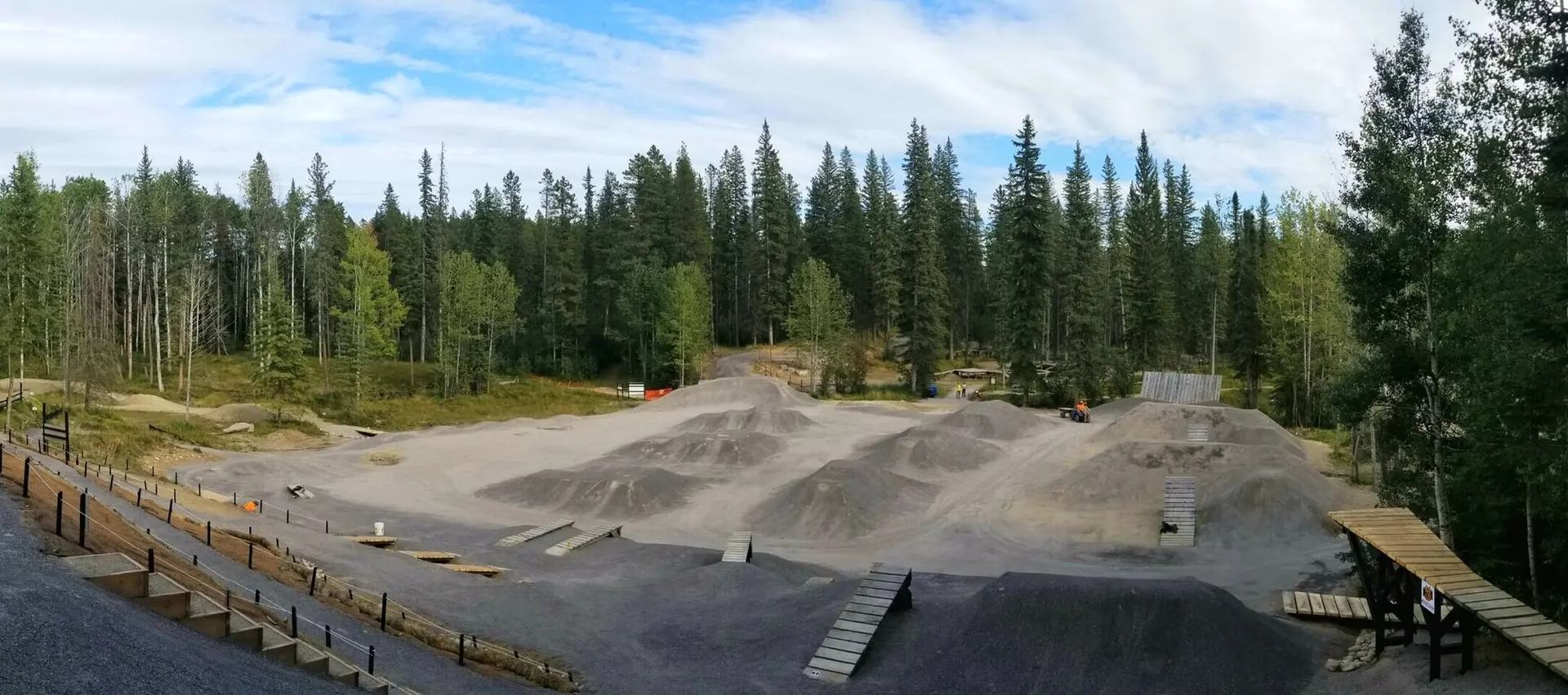 Gravel bike park with dirt jumps and wooden ramps surrounded by dense evergreen forest.