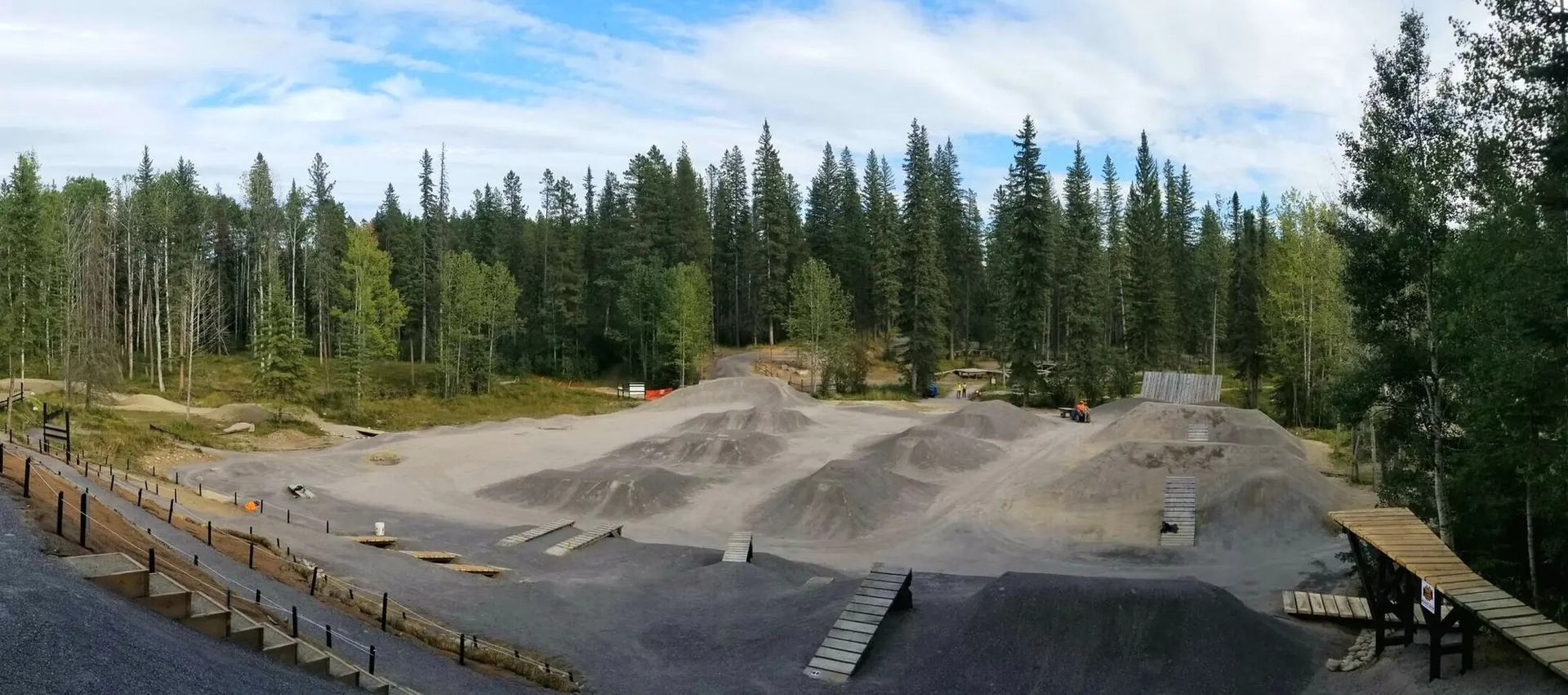 Gravel bike park with dirt jumps and wooden ramps surrounded by dense evergreen forest.