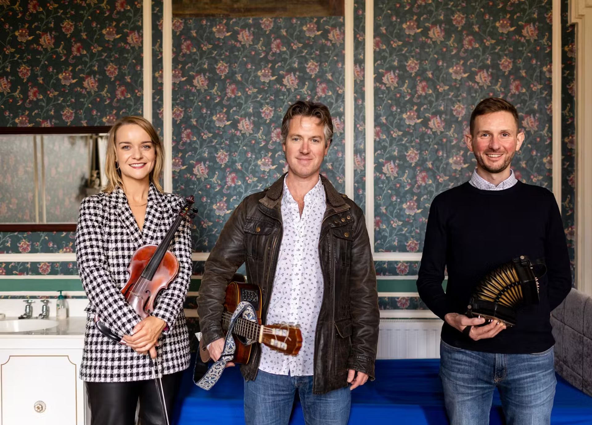 Three musicians with violin, accordion, and concertina in front of floral wallpaper.