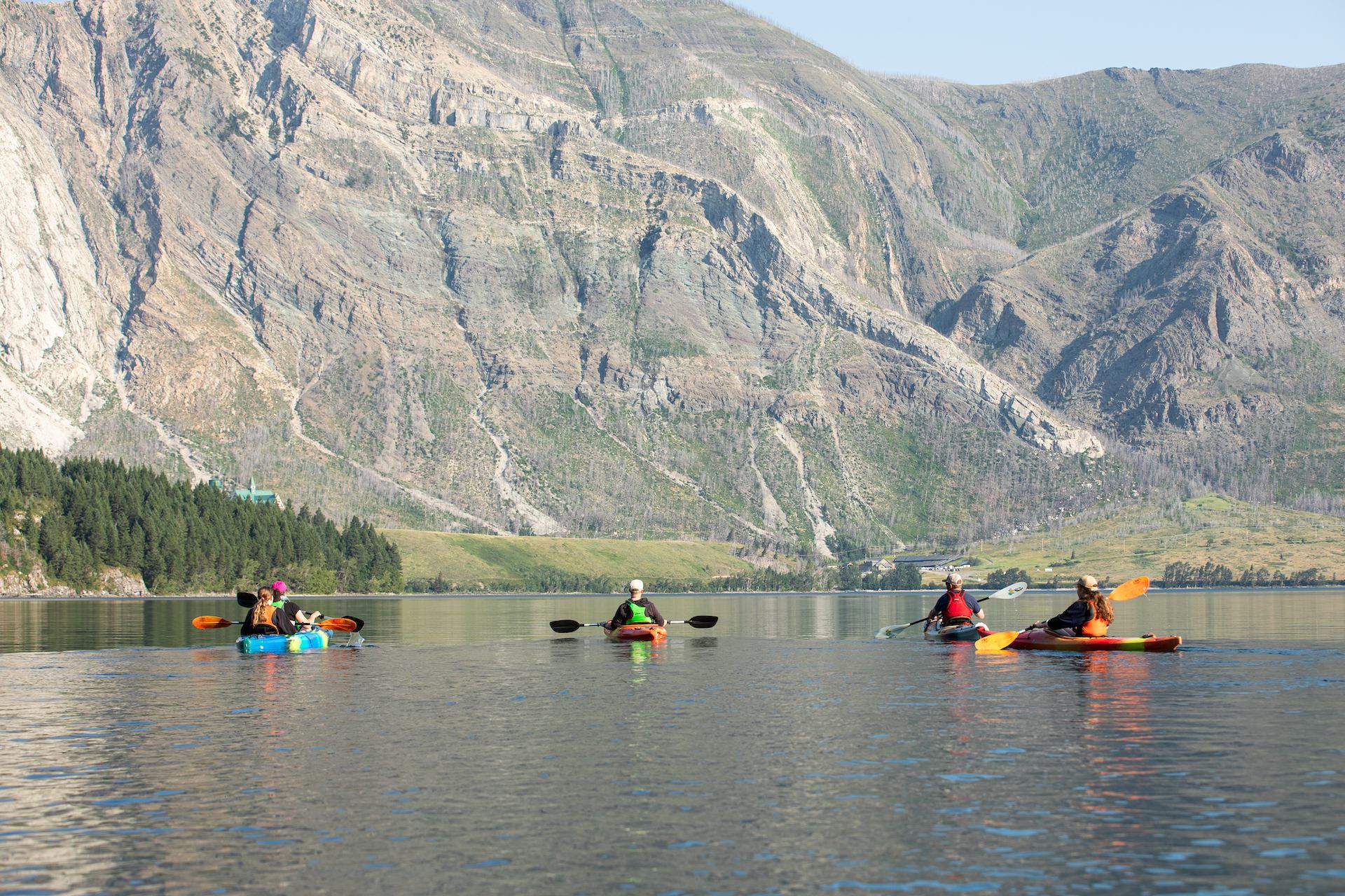 Kayakers paddle across calm water with rocky mountains in the background.