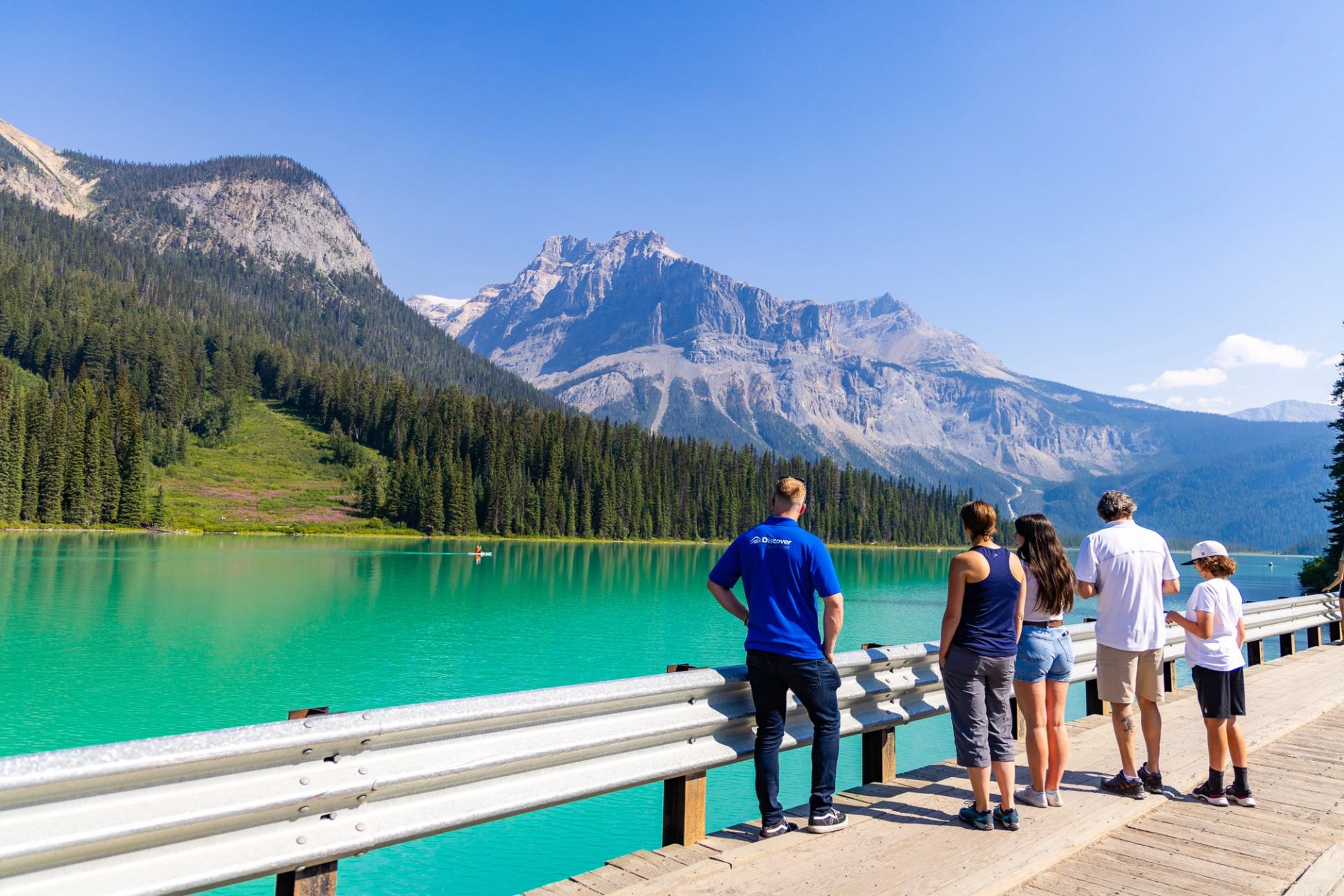 People stand on a wooden boardwalk overlooking turquoise lake and forested mountain range.