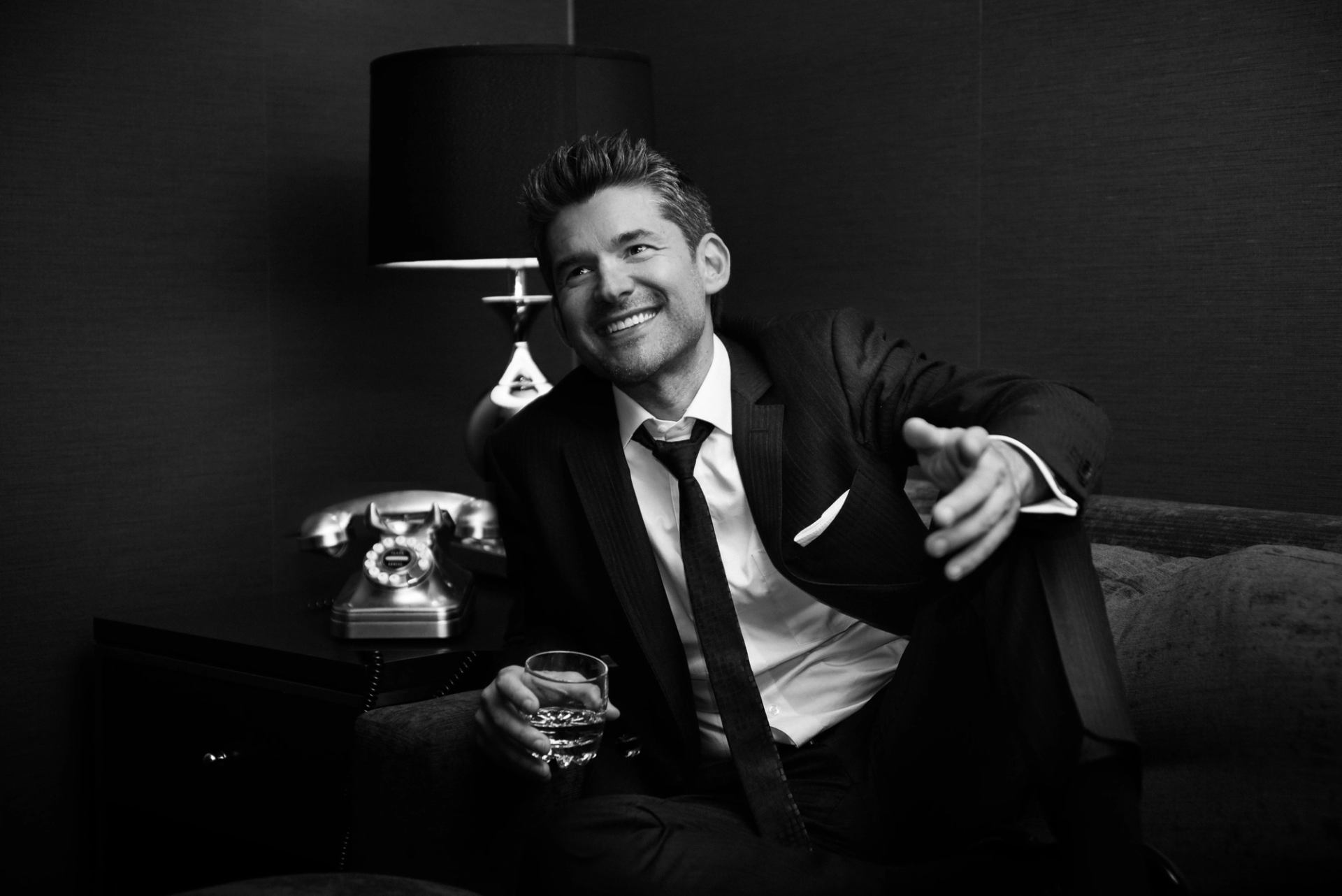 Man in suit sits on sofa, holding drink beside retro telephone and lamp.