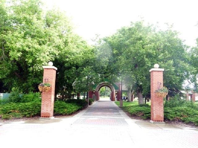 Brick pillars with hanging baskets frame a paved walkway under trees to an arch.