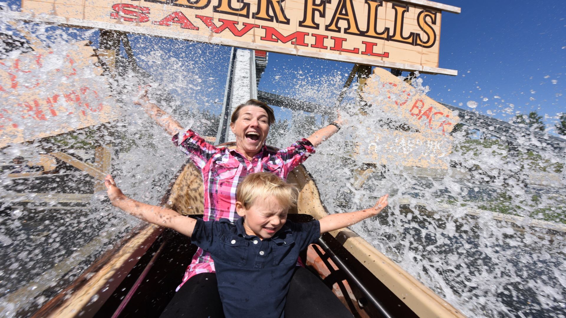 People ride a log flume as water splashes up near Timber Falls Sawmill sign.