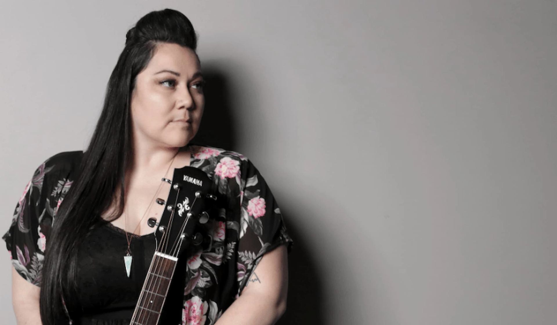 Person holding a Yamaha acoustic guitar, wearing floral top, standing against plain wall.