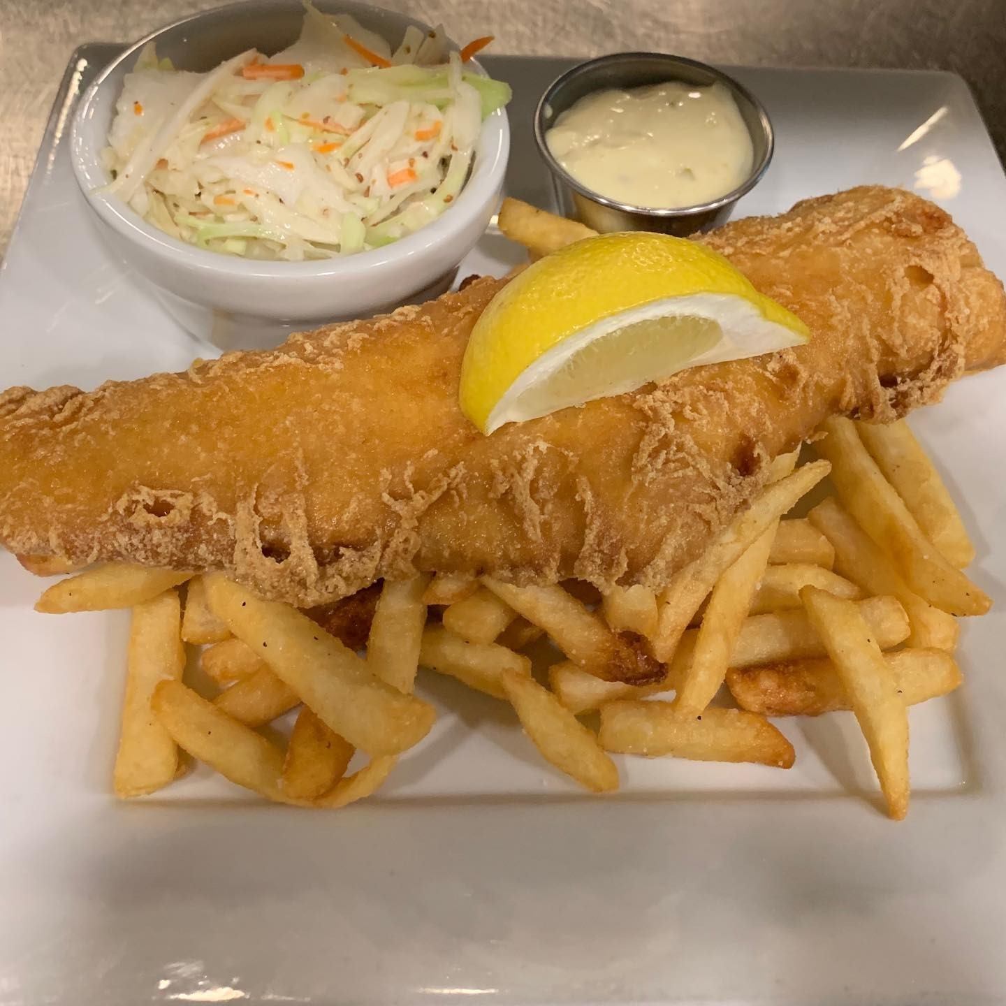 Fish and chips with lemon wedge, coleslaw and dipping sauce on white plate.