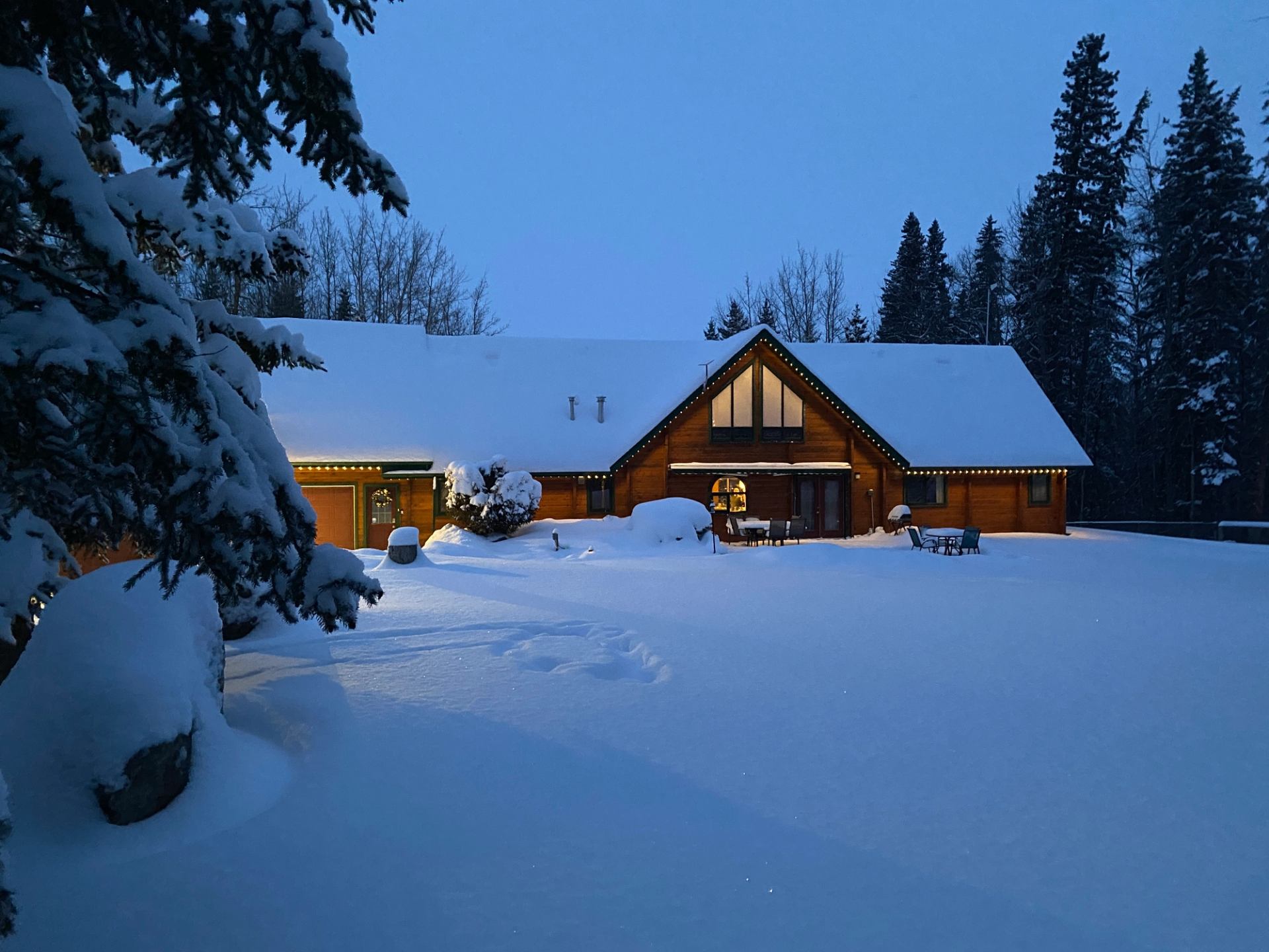 Snow-covered cabin with warm lights, surrounded by trees and deep snowdrifts at dusk.