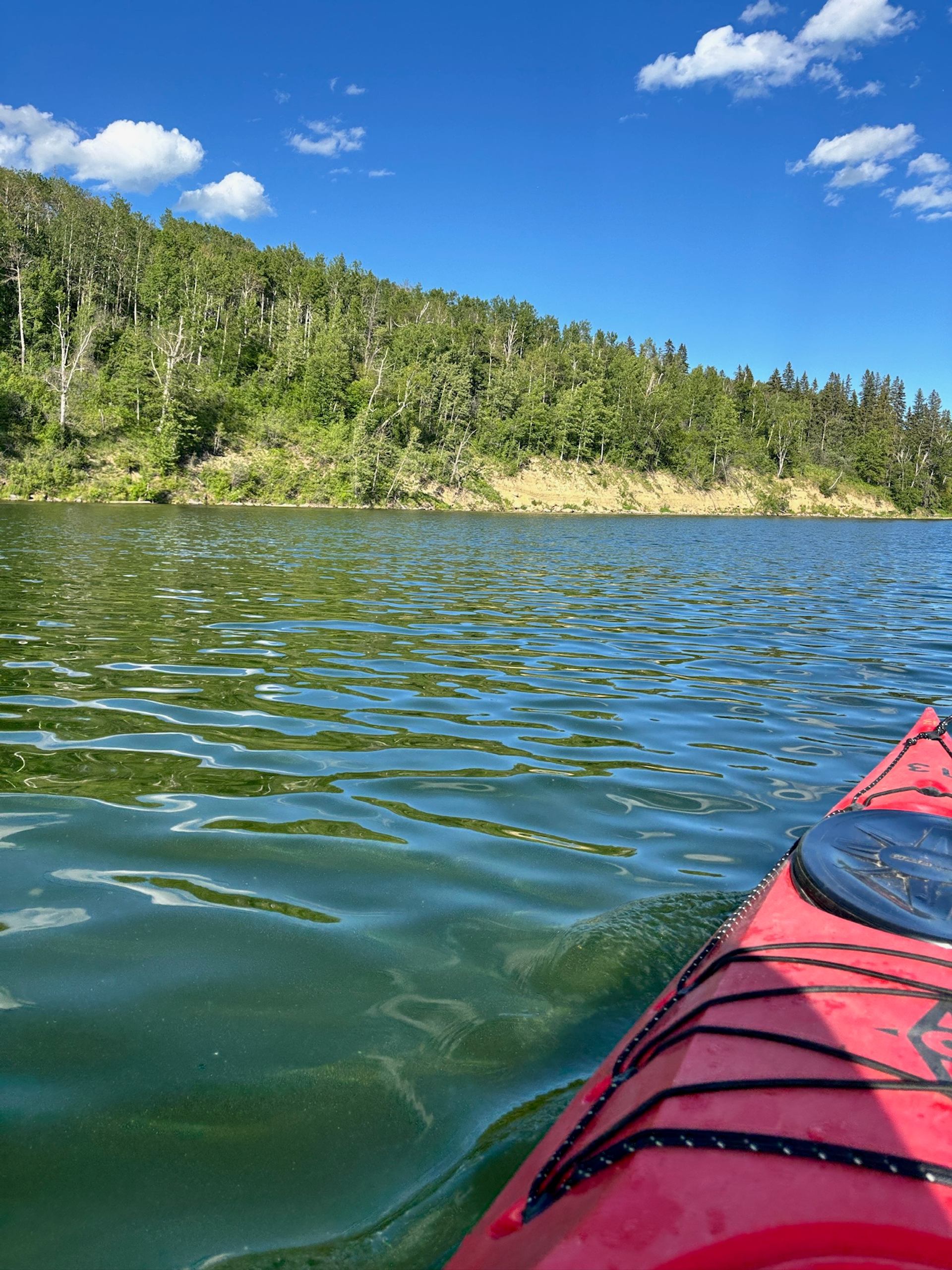 Red kayak bow on rippled lake, forested shoreline and blue sky with clouds.