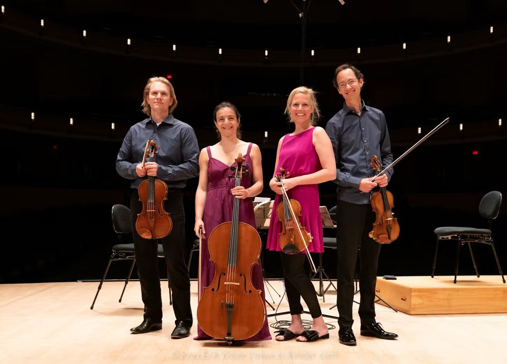 Four string musicians on stage in auditorium with violins, viola, and cello.