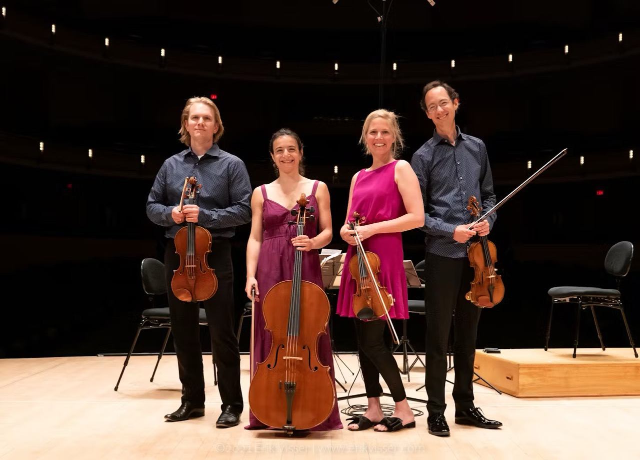 Four string musicians on stage in auditorium with violins, viola, and cello.