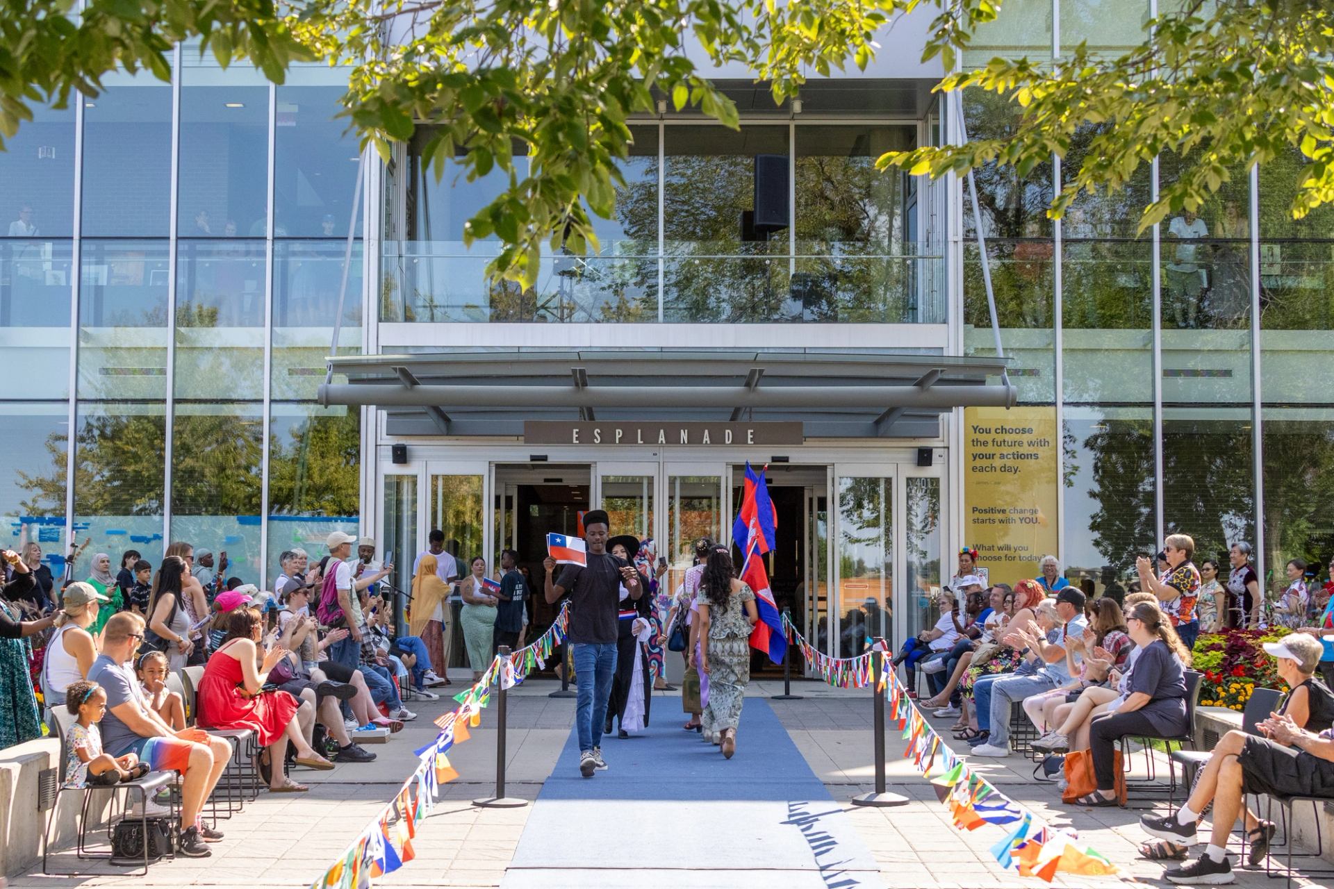 Outdoor crowd watches people carrying colourful flags walk a blue carpet into Esplanade building.