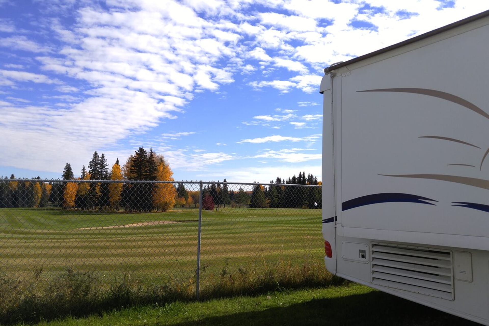 RV parked beside a chain‑link fence overlooking a large grassy field and trees.