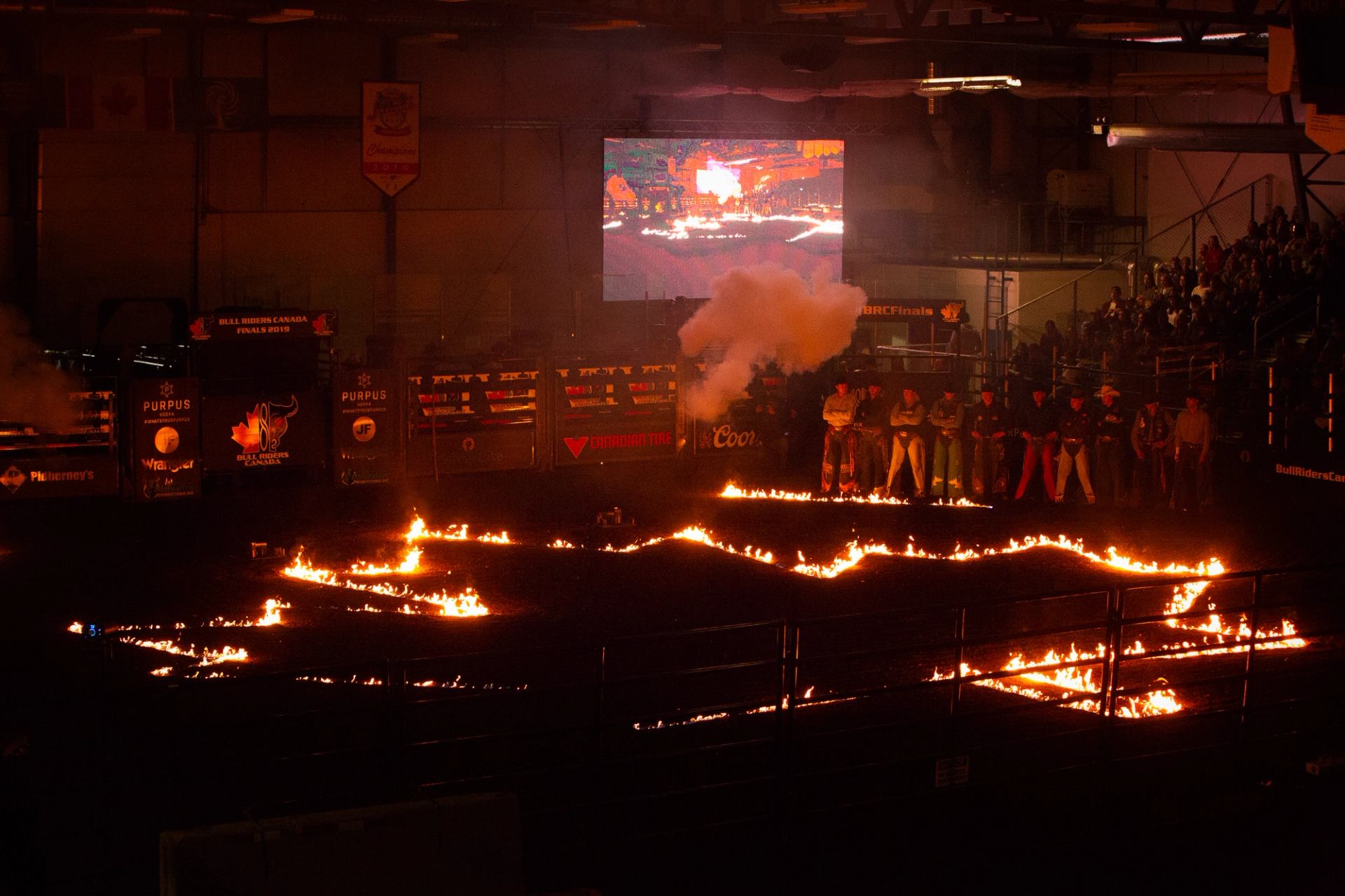 Indoor rodeo arena with flaming patterns on dirt and riders standing near smoke.