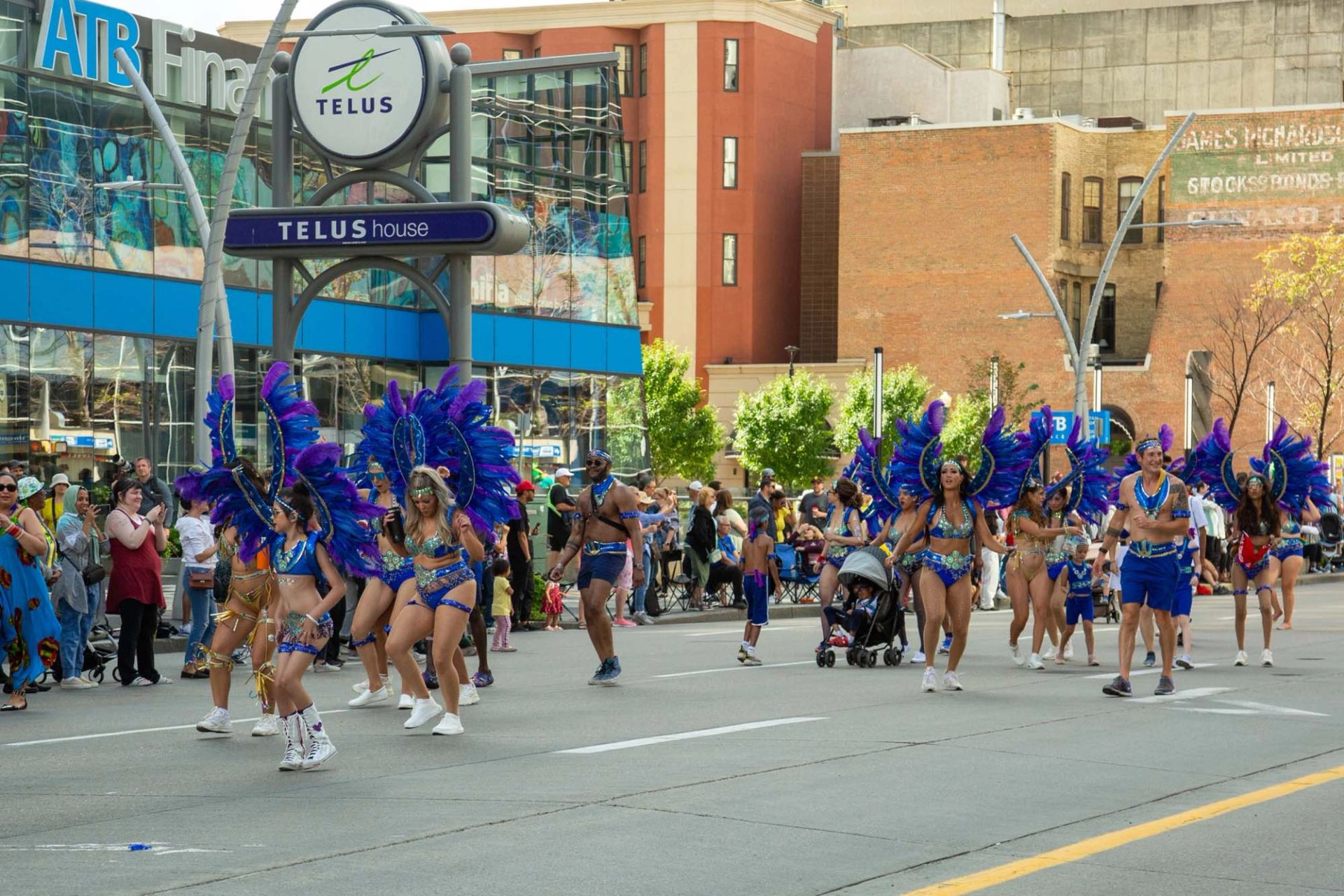 Parade performers in blue feathered costumes dance along a downtown street past TELUS building.
