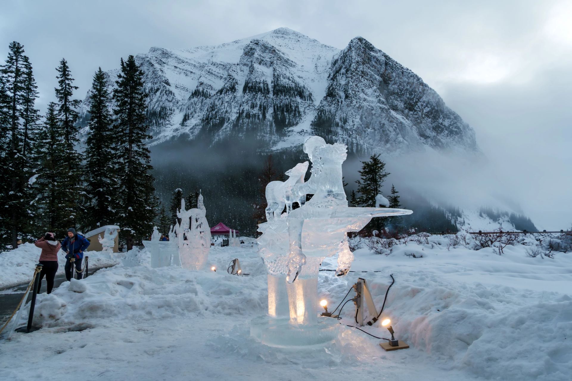 Ice sculptures displayed in a snowy mountain setting with mist and tall pine trees.