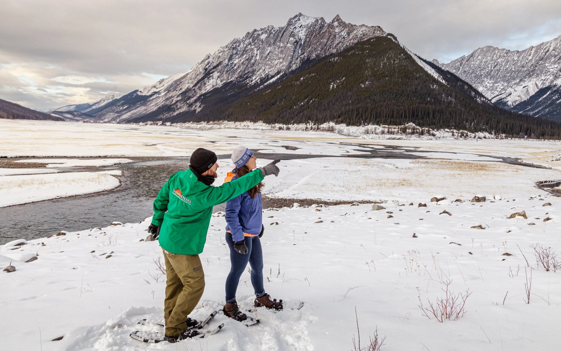 Two travelers snowshoe beside a frozen river, pointing toward snow-covered mountains in the Canadian Rockies.