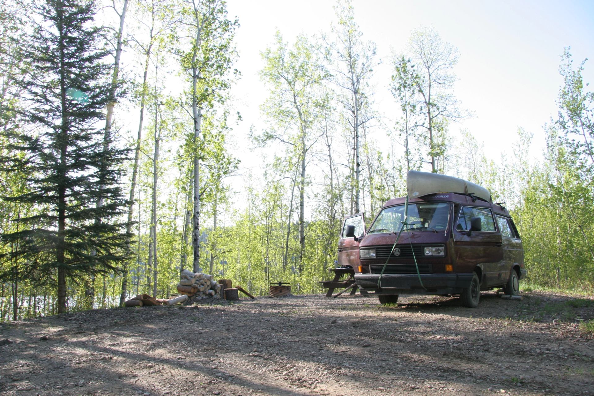 Red camper van with canoe parked at forest campsite beside stacked firewood.