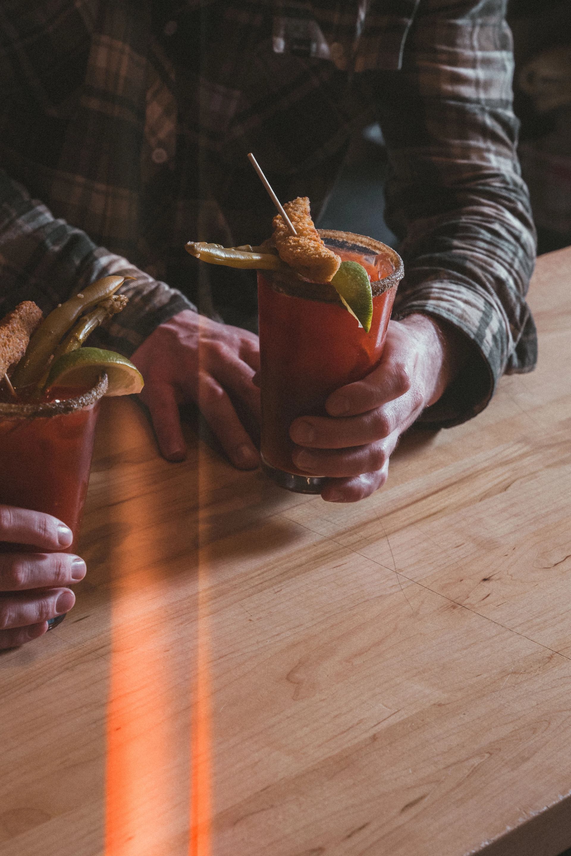 A person in a plaid shirt holds two Bloody Mary cocktails garnished with pickles and limes on a wooden bar.