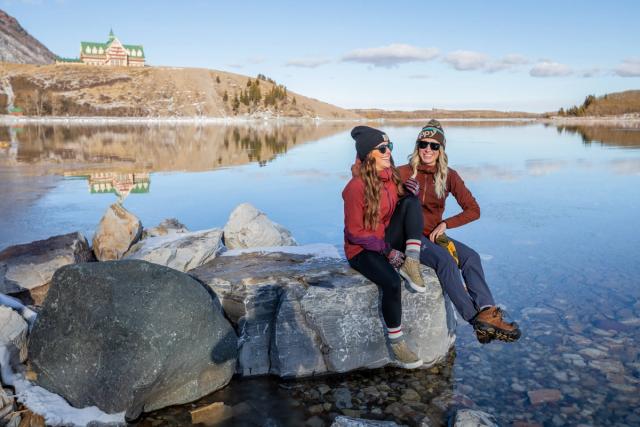 Two girls sit and talk on a rock in the water with the Prince of Wales Hotel in the background.