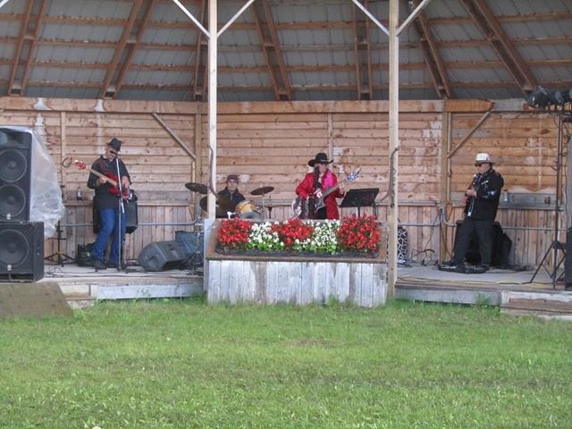 Band performs on outdoor wooden stage with instruments, speakers and red flower planter.
