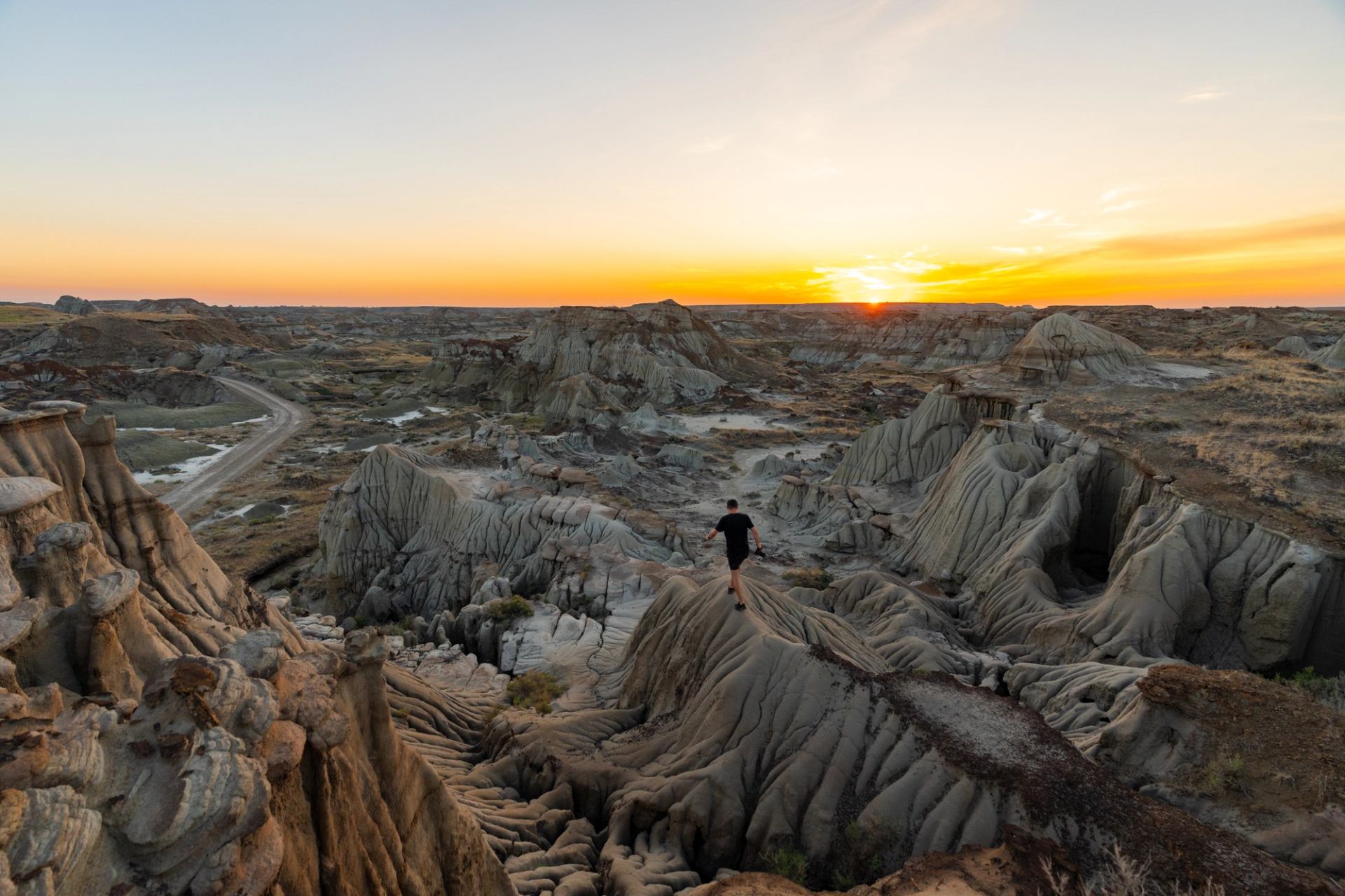 A person walking along rugged, eroded rock formations in the badlands during sunset, with winding dirt paths, layered hills, and warm orange light illuminating the landscape.
