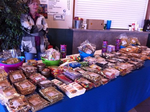 Smoky Lake Farmers Market features a vendor table covered with a wide variety of packaged baked goods, snacks, and handmade treats neatly arranged, with a vendor standing behind the display in an indoor market setting.