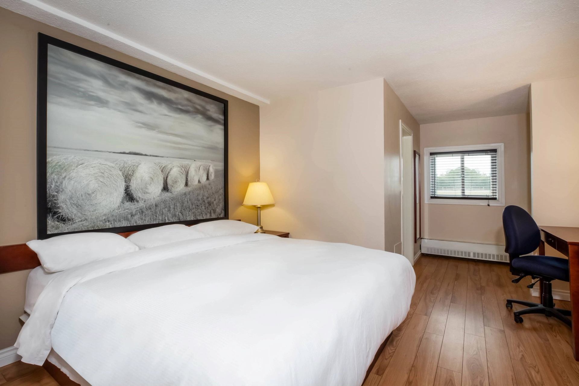A hotel room with a white bedspread, framed artwork of hay bales, a desk with a chair, and a window. The room has light wooden flooring and soft lighting.