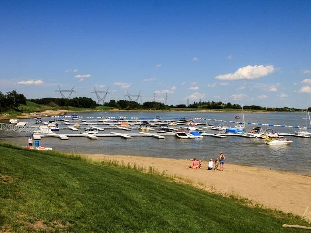 Lakefront view with boats, beachgoers, grassy area, and blue sky with clouds.