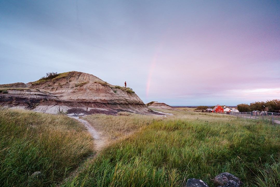 Rocky hill beside a dirt path and red buildings under a pink sky.