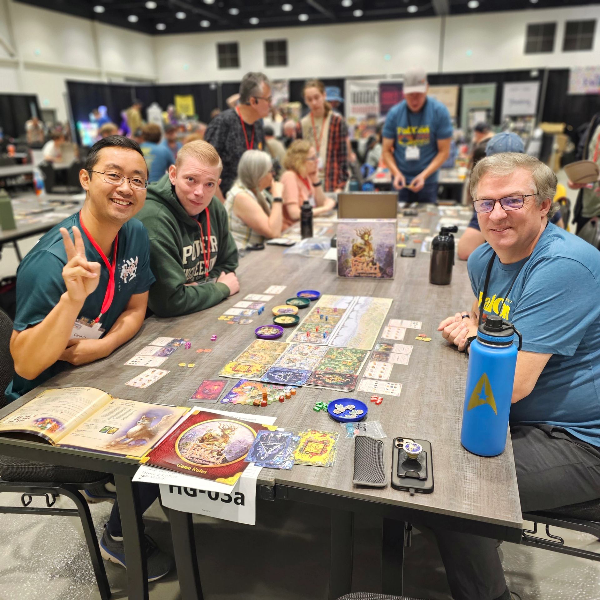 Board game players sit at a convention table covered with colourful cards and components.