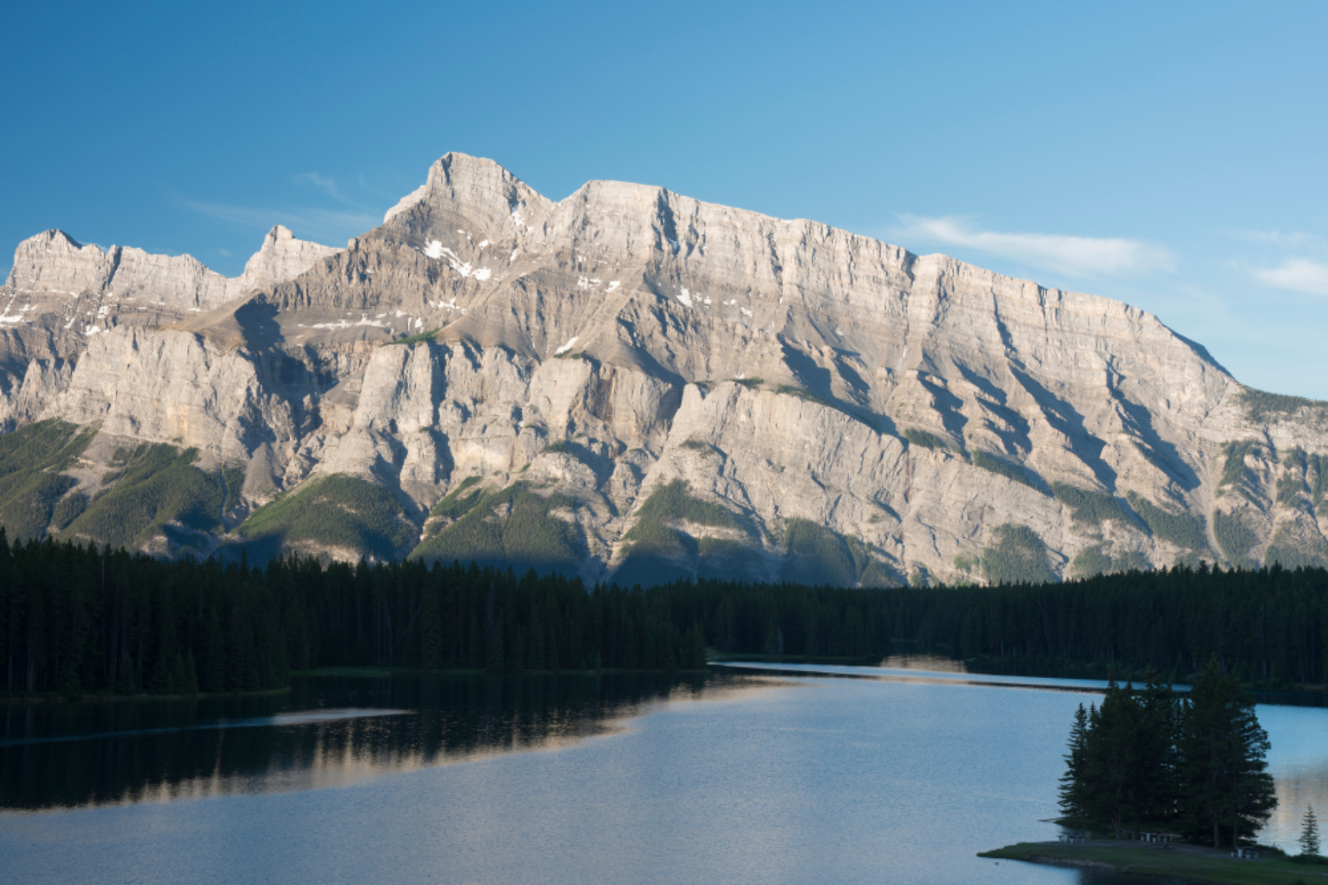 Rocky mountain rising above a calm lake with forest along the shoreline on a clear day.